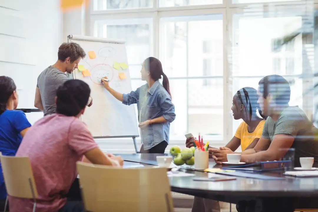 A diverse team round a meeting table while one colleague writes on a flip chart